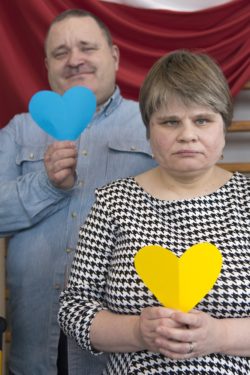 Kobieta trzymająca w dłoniach żółte serce. Za nią styłu mężczyna trzymający niebieskie serce.