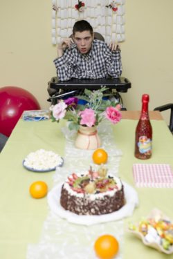 Siedzący na wózku inwalidzkim chłopiec za stołe na którym stoją tort, owoce, flakon z kwiatami i słodycze.