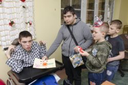 Trzech chłopców wręczających upominki chłopcu siedzącemu na wózku inwalidzkim.