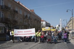 Grupa osób maszerująca ulicą, niosąca baner z napisem Marsz Godności Osób Niepełnosprawnych.