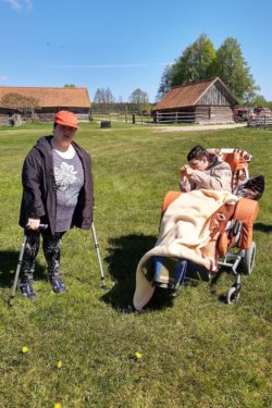 Dwie kobiet, jedna wsparta na kulach, druga na wózku inwalidzkim na tle wiejskich zabudowań.