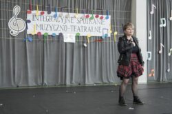 Kobieta z mikrofonem w ręku stojąca na dużej sali. Za nią w tle na ścianie napis: integracyjne spotkania muzyczno-teatralne.