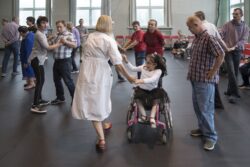 Kobieta tańcząca z dziewczynką na wózku inwalidzkim. W tle inne tańczące osoby.
