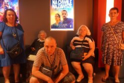Grupa osób stojąca w kinie przy plakacie filmowym.