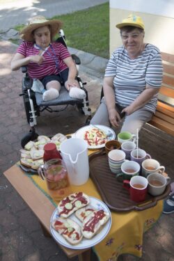 Kobieta na wózku inwalidzkim i kobieta siedząca na ławce przy stoliku na którym stoi tależ z kanapkami i taca z napojami.