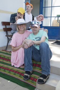 Kobieta i mężczyzna w letnich kapeluszach siedzący na rozłożonym na schodach kocu. W tle za nimi trzy osoby siedzące na ławce.