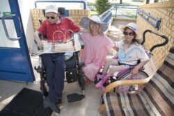 Mężczyzna na wózku inwalidzkim i dziewczynka na wózku inwalidzkim, pomiędzu nimi przykucnięta kobieta w letnim kapeluszu.