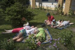 Grupa osób leżąca na kocu rozłożonym na trawniku wśród kwiatów.
