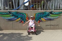 Dziewczynka na wózku inwalidzkim przed muralem przedstawiającym skrzydła.