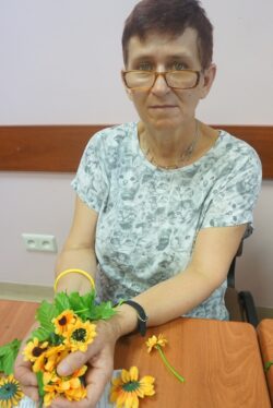 Siedząca przy stole kobieta trzyma w dłoniach kolorowe kwiaty.