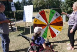 Dziewczynka na wózku inwalidzkim przed dużym kolorowym kołem z numerami. Po jej obu stronach stojące dwie inne osoby.