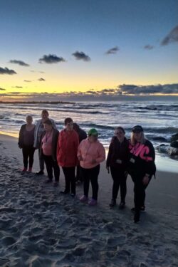 Grupa kobiet stojąca na plaży na tle morza o zachodzie słońca.