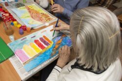 Siedząca przy stole kobieta malująca farbami obrazek.