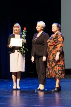 Trzy kobiety stojące na scenie. Jedna z kobiet trzyma w rękach kwiat i dyplom.
