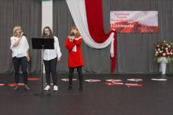 Trzy kobiety z mikrofonami w rękach stojące na dużej sali na tle biało-czerwonych dekoracji.