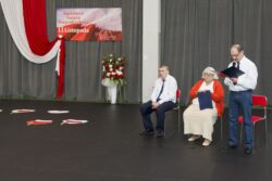 Mężczyzna z teczką i mikrofonem w dłoni stojący na dużej sali. Po lewej stronie na krzesłąch siedzą mężczyzna i kobieta. W tle biało-czerwone dekoracje.