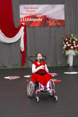 Dziewczynka na wózku inwalidzkim z mikrofonem w ręku na tle biało-czerwonych dekoracji.