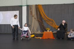 Stojący na dużej sali mężczyzna trzymający za rękę dziewczynkę na wózku inwalidzkim. Obok nich siedzący w fotelu przy stoliku ksiądz.