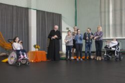 Grupa osób stojąca na dużej sali i dziewczynka na wózku inwalidzkim z mikrofonem w ręku.