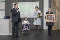 Mężczyzna z mikrofonem w ręku, dziewczynka na wózku inwalidzkim i dwie kobiety stojący na dużej sali. Jedna z kobiet trzyma obraz druga bukiet kwiatów.