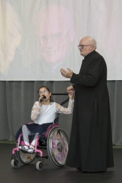 Ksiądz i dziewczynka na wózku inwalidzkim z mikrofonem w dłoni.