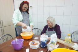 Dwie kobiety przy zastawionym kuchennymi produktami i naczyniami stole. Jedna z kobiet za pomocą miksera miesza zawartość miski.