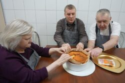 Siedząca przy stole kobieta i dwaj mężczyźni trzymający w rękach naczynie z ciastem.