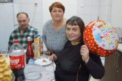 Mężczyzna i dwie kobiety siedzący przy zastawionym słodyczami i napojami stole. Jedna z kobiet trzyma w rękach kolorowy balonik.
