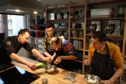 Czterech mężczyzn przygotowujących potrawę przy dużym stole kuchennym.