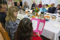 Grupa siedzących wokół zastawionego naczyniami i słodyczami stołu osób. Jedna z kobiet trzyma przed sobą w rękach kartkę z tekstem.