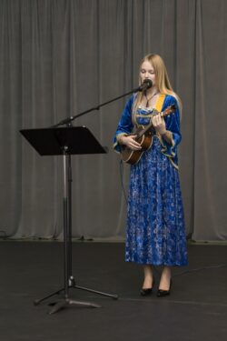 Kobieta z ukulele w rękach stojąca przed mikrofonem i pulpitem.