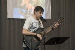 Mężczyzna z gitarą w rękach przed mikrofonem i pulpitem. W tle kolorowy obrazek.