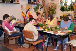 Pięć kobiet siedzących przy stoliku na którym leżą różne przedmioty do wykonywania stroików. W tle kolorowe stroiki.