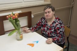 Siedząca przy stole kobieta przed którą leży kolorowy obrazek i stoją kwiaty w naczyniu.