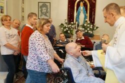 Grupa osób w kaplicy. Na pierwszym planie mężczyzna na wózku inwalidzkim przyjmujący komunię.