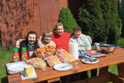 Cztery kobiety siedzące na ławce przy stole zastawionym różnymi naczyniami i produktami żywnościowymi. 