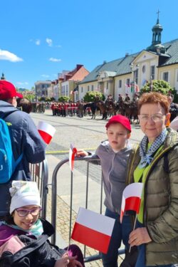 Kobieta i dwoje dzieci z biało czerwonymi chorągiewkami stający przy barierce na dużym placu. W tle budynki i ustawieni w szeregi żołnierze.