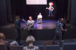 Grupa osób tańcząca przed sceną na której jest mężczyzna na wózku inwalidzkim.