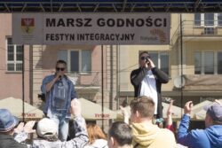 Dwaj mężczyźni z mikrofonami w rękach przed duża grupą osób. Za nimi w tle widoczny napis: Marsz Godności, festyn integracyjny.