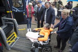 Kobieta wprowadzająca wózek inwalidzki na podnośnik auta. Na wózku inwalidzkim siedzi kobieta. W tle inne osoby.