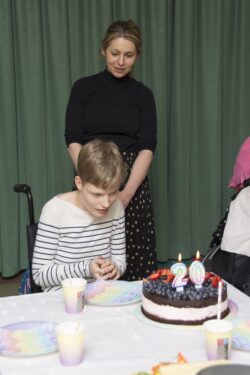Dziewczynka siedząca przy stole i stojąca za nią kobieta. Na stole stoi tort i plastikowe naczynia.