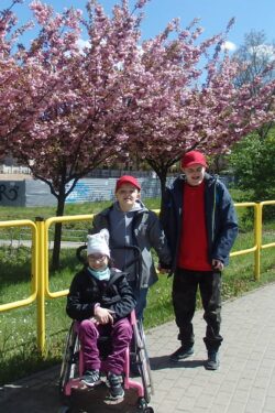 Dziewczynka na wózku inwalidzkim i dwaj chłopcy na tle kwitnących drzew.