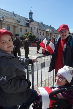 Dziewczynka na wózku inwalidzkim i dwaj chłopcy trzymający w rękach biało czerwone chorągiewki stojący przy barierkach na dużym placu. W tle budynki i żołnierze.