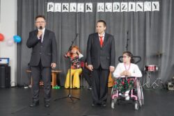 Mężczyzna w garniturze z mikrofonem w ręku i stojący obok niego chłopiec w garniturze i dziewczynka na wózku inwalidzkim. Za nimi w tle napis: piknik rodzinny.