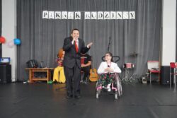Chłopak w garniturze i dziewczynka na wózku inwalidzkim na dużej sali. Za nimi w tle napis: piknik rodzinny.