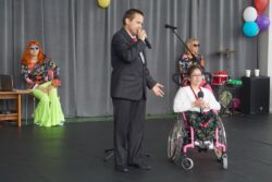 Chłopiec z mikrofonem w ręku i dziewczynka na wózku inwalidzkim na dużej sali. W tle dwie inne osoby.