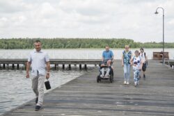 Grupa osób idąca drewnianym pomostem na jeziorze.