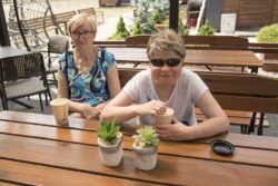 Dwie kobiety siedzące przy stoliku na którym stoją kwiaty i kubki z kawą.