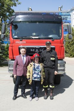 Dwaj mężczyźni i kobieta w stroju strażaka stojący przed wozem strażackim.