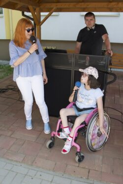Kobieta i dziewczynka na wózku inwalidzkim z mikrofonami w ręku stojące pod altaną. W tle mężczyzna.